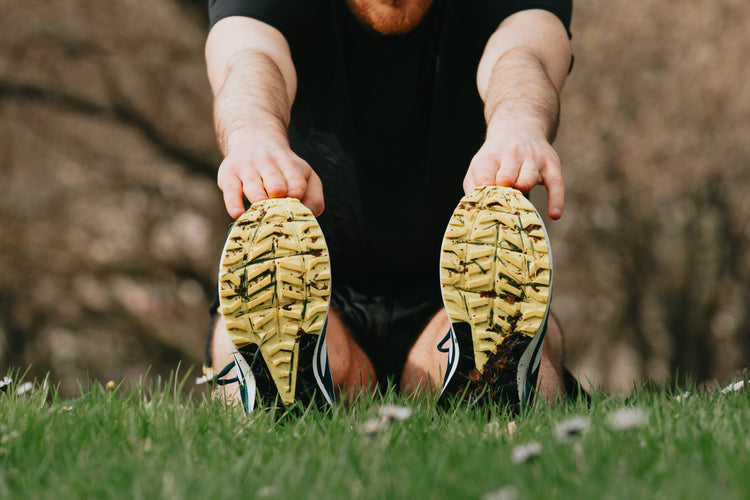 Person stretching with hands reaching towards their shoes in a grassy outdoor setting.